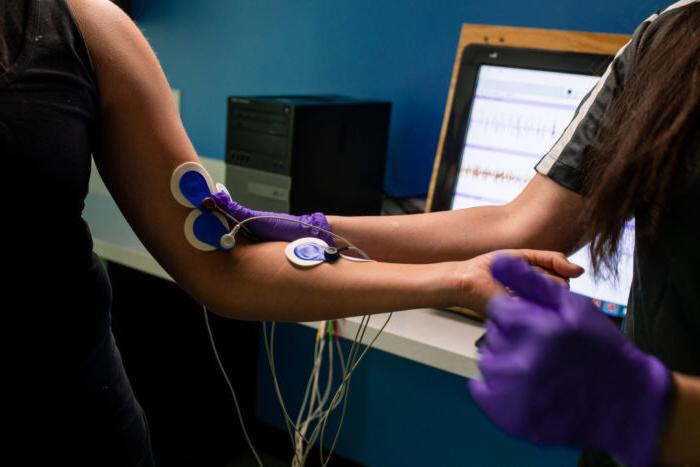 A person wearing purple gloves attaches electrodes to another person's arm while monitoring data on a computer screen in a medical or research setting.