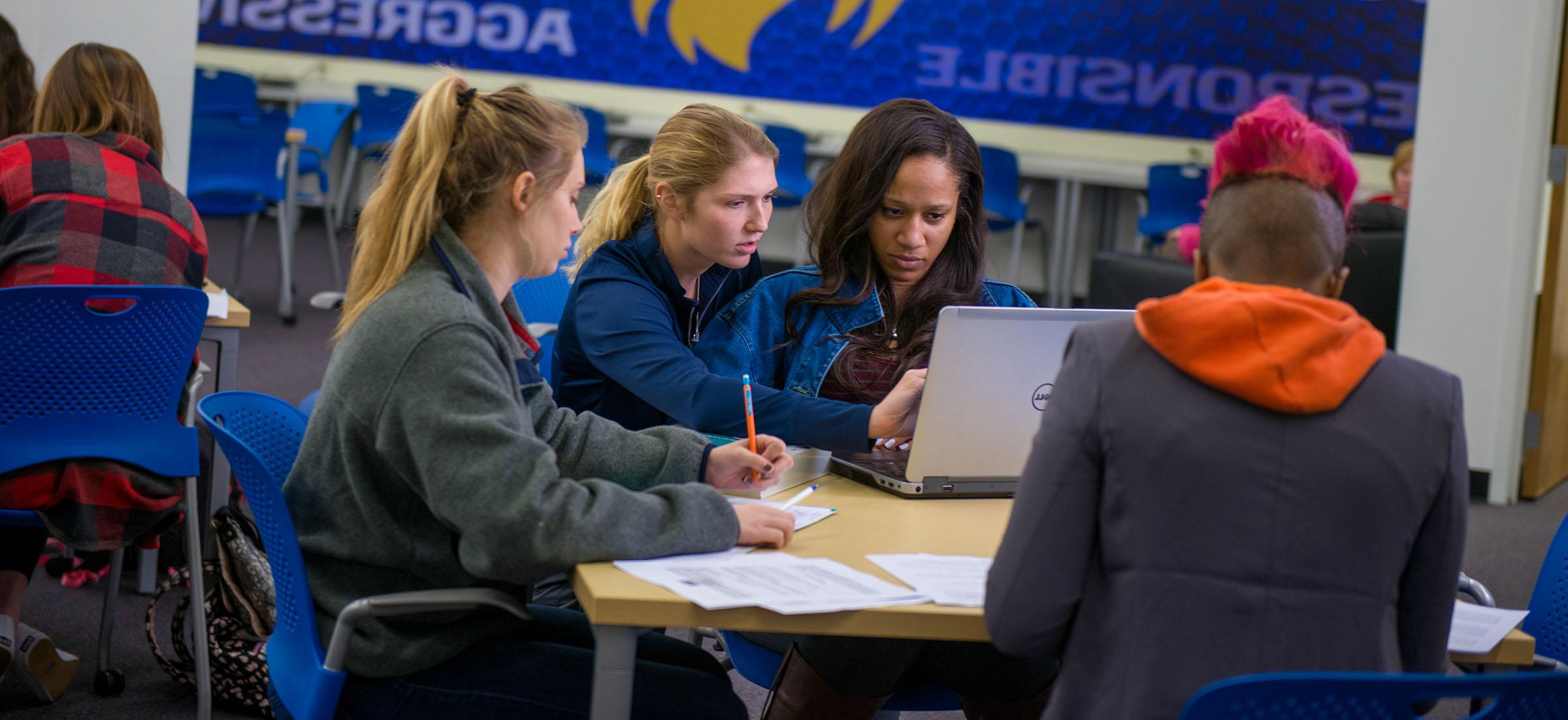 A group of student completing an assignment together. They are at a desk with other desks around them. In the background there is blue with words on it.