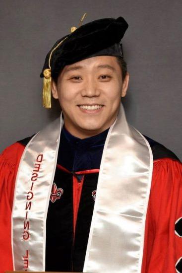 A graduate dressed in an academic gown and cap smiles warmly at the camera. He wears a black academic hat and a white stole with red lettering that reads "Designing Leaders.