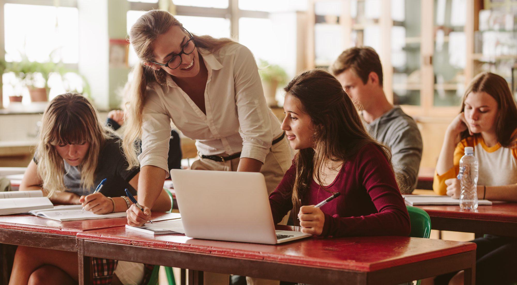 A female teacher helping a female student with a few students in the background in a classroom