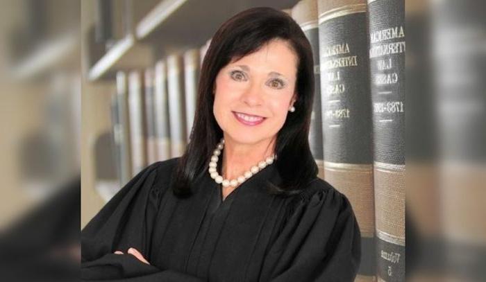 A person in a judge's robe leaning against bookshelves.