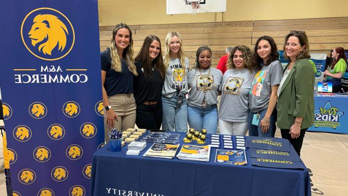 Seven people stand shoulder-to-shoulder in front of a table with a blue cloth cover and promotional items on top. Next to them is a blue, vertical banner with the A&M-Commerce logo and numerous smaller lion head logos below.